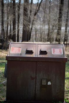 Large rusted receptacle for trash and aluminum and plastic recyclables in front of a lake