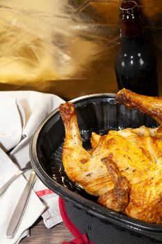 Whole roasted chicken in a black carryout container and a bottle of beer next to a grey napkin and silverware