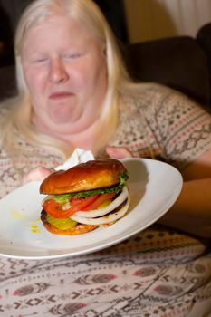 Woman sadly eating a burger with lettuce, tomato, white onion, and pickles on a white plate