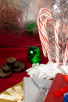 Red striped candy canes hanging on a crystal vase next to white bows, silver, red, and blue presents, and Christmas chocolate on a red tabletop