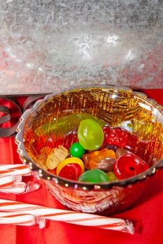 Multicolored candies in a small bowl next to candy canes on a red tabletop with a tin sleigh in the background