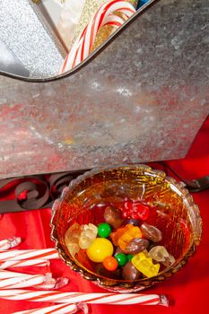 Multicolored candies in a small candy bowl next to candy canes and a tin sleigh on a red tabletop