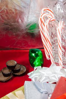 Candy canes on a glass vase with chocolates, green ribbon paper and gold, silver, and red presents on a red tabletop