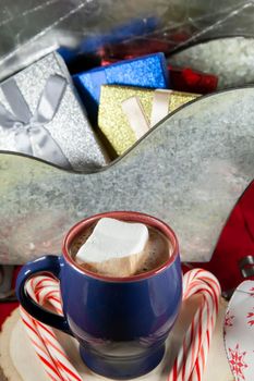 Blue and red mug filled with hot chocolate and a marshmallow, candy canes on a wooden stump, with a tin sleigh filled with silver, gold, blue, and red gifts in the background