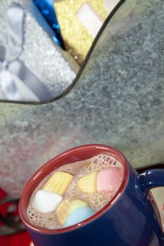 Blue and red mug with multicolored marshmallows next to a tin sleigh filled iwth silver, blue, and gold presents in the background