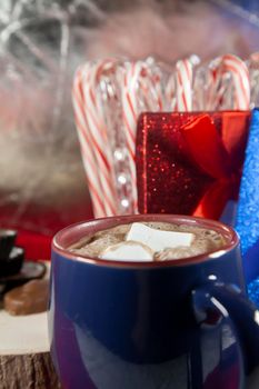 Blue and red mug filled with hot chocolate and two marshmallows, with chocolates on a wooden stump and candy canes and red and blue presents in the background