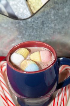Blue and red mug with multicolored marshmallows on a wooden surface with candy canes, next to a tin background