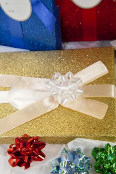 Golden gift box topped with a white bow in front of blue and red gift boxes and next to red, blue, and green bows