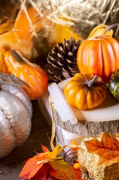 Small orange pumpkins, orange squash, pinecones, a green squash, and glue sticks for crafting on wood stacked on leaf-covered hay next to a silver pumpkin with a golden background