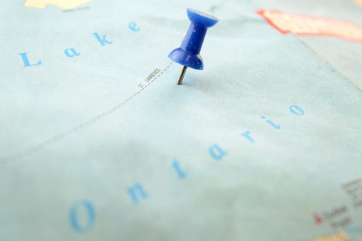 A blue pin point pressed into a map detailing Lake Ontario which is one of the Great Lakes that border Canada and the United States of America.