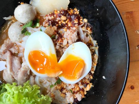 Thai spicy pork noodle soup with coconut milk, vegetables herbs and onsen tamago eggs  in black bowl with sesame seeds and peanut on wooden table, view from above