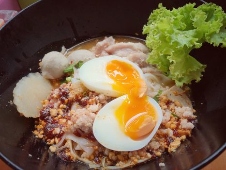 Thai spicy pork noodle soup with coconut milk, vegetables herbs and onsen tamago eggs  in black bowl with sesame seeds and peanut on wooden table, view from above