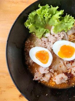 Thai spicy pork noodle soup with coconut milk, vegetables herbs and onsen tamago eggs  in black bowl with sesame seeds and peanut on wooden table, view from above