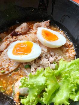 Thai spicy pork noodle soup with coconut milk, vegetables herbs and onsen tamago eggs  in black bowl with sesame seeds and peanut on wooden table, view from above