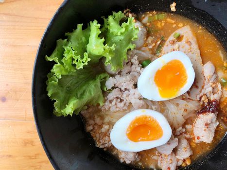 Thai spicy pork noodle soup with coconut milk, vegetables herbs and onsen tamago eggs  in black bowl with sesame seeds and peanut on wooden table, view from above