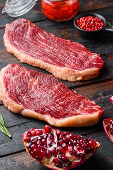 Picanha raw organic beef steaks with seasoning, rosemary, tpmators, spicy chili oil and pomegranates over old dark wood background, side view selective focus