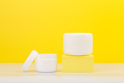 Two cream jar with the bigger one on podium on white glossy table against yellow background. Minimalistic concept of daily beauty routine and skin treatment