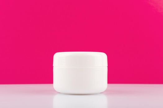 Minimalistic still life with white cream jar on white glossy table against pink background with copy space. Concept of beauty and skincare or anti aging treatment