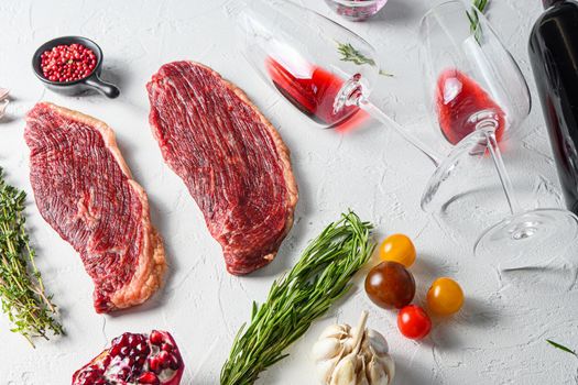 Picanha organic beef steaks with rosemary, peppercorns, pomegranate, near red wine in glasses and bottle over white textured background, side view.