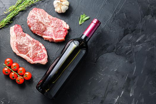 Raw top blade steak cuts, with red wine bottle and glasses, herbs and seasonings on black textured background side view with space for text