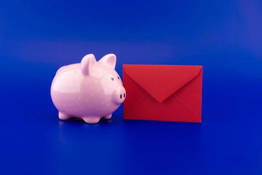 Cute pink piggy bank and red envelope over blue background with copy space
