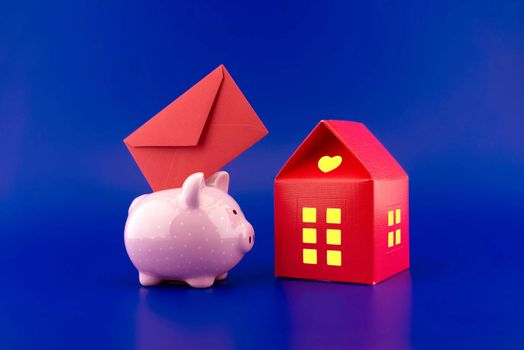 Red envelope being inserted into a cute pink piggy bank next to a small red house of paper with warm light in windows over festive blue background with copy space