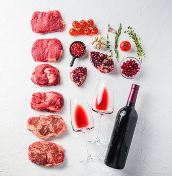 Rraw beef steaks set top blade, rump, chuck eye roll with herbs, red wine bottle and glasses over white concrete background, top view