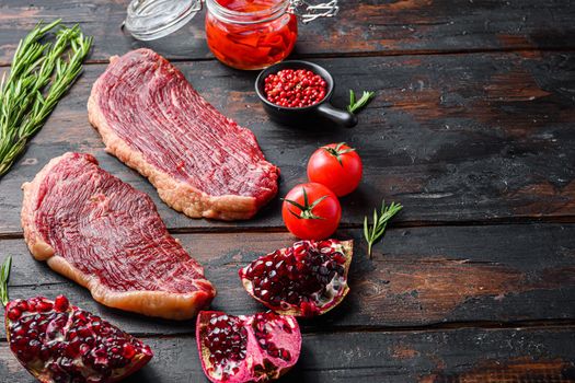 Picanha raw organic beef steaks with seasoning, rosemary, tpmators, spicy chili oil and pomegranates over old dark wood background, side view space for text