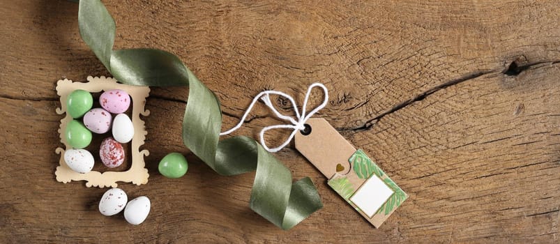 Easter background with colourful eggs, ribbon, gift tag on old wooden background. Top view with copy space. Rustic Easter concept