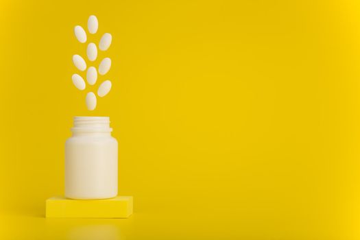 Still life with opened medication bottle on podium with white oval pills jumping out against yellow background with copy space. Concept of vitamins for kids or wellbeing and pharmacy