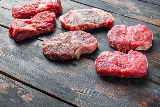Variety of organic raw beef steakes rump, chuck roll and top blade cut on old rustic dark wooden background, side view with space for text