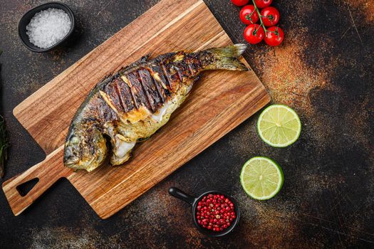 Grilled sea bream or dorado raw fish on chopping board with ingredients over old metall dark background, top view with space for text