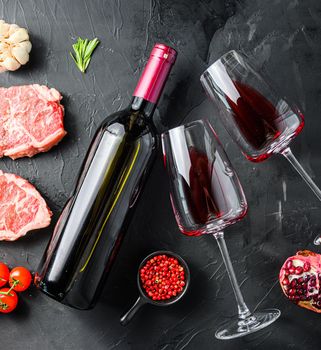 Red wine bottle with two glasses near top blade steaks with herbs seasoning over black textured background top view square