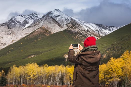 Pretty woman takes pictures on the smartphone of the yellow larches and snowy mountains at Kananaskis National Park, Canada.