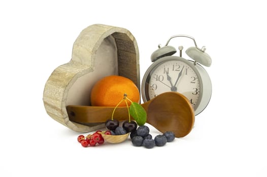 Culinary still life with old-fashioned alarm clock, wooden spoons, cherries and orange, red currants and blueberries over an off white background with copy space