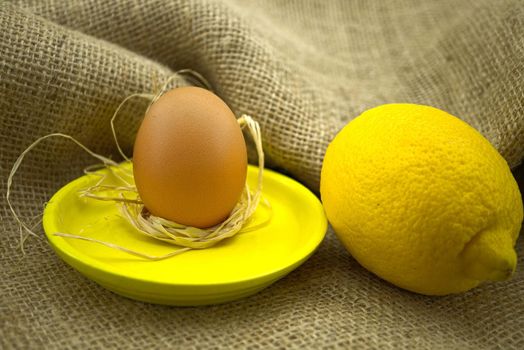 Brown egg on bright yellow plate near yellow lemon over rustic hessian sack background. Organic farming and color trends concept with copy space
