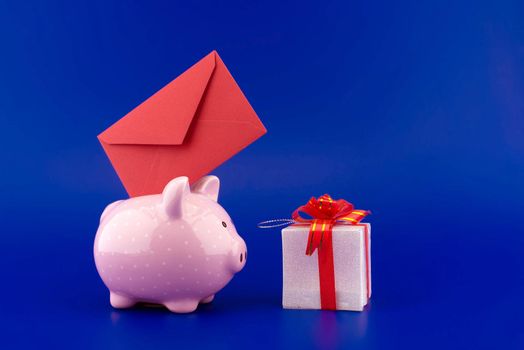 Festive red envelope being inserted into a pink piggy bank next to a present box over blue background with copy space