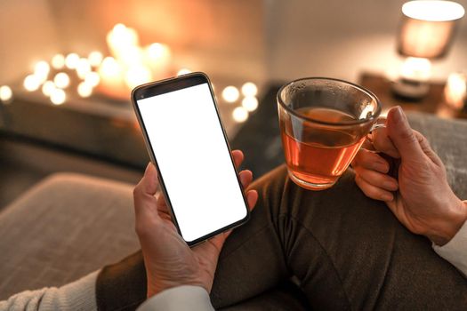 New reading technology using smartphone to enjoying preferred ebook: close up of unrecognizable woman sitting on sofa holding a cup of tea in one hand and big display cell phone with blank white screen