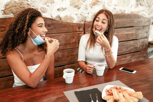 Two young beautiful mixed-race women sitting outdoors on rustic wooden table wearing lowered protective mask eating some pastries smiling looking at each other with coffee cup and smartphone