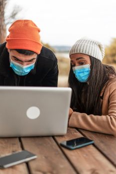 Young multiracial couple of friends using laptop outdoor wearing protective mask and winter clothes. Hispanic girl sitting at a wooden table in pine forest browsing on internet during winter vacation