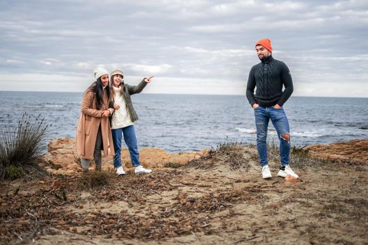 Two girls make fun of a guy outdoors by pointing that he has an amused expression. Three best multiracial friends having fun outdoor joking to each other in sea resort during winter vacation
