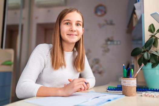 Beautiful happy schoolgirl studying at home doing school homework. Training books and notebook on the table. Distance learning online education