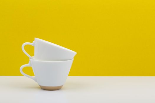 Minimalistic still life with two white coffee cups on in another on white table against yellow background with copy space. 