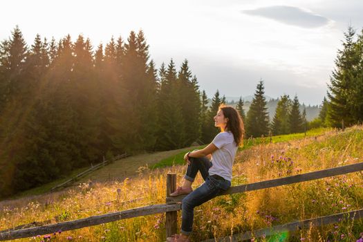 The girl enjoy sunset sitting on wooden fence on highlands field next to forest.