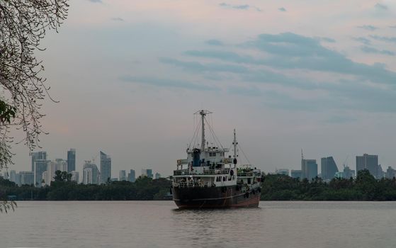 Bangkok, Thailand - 04 Jan 2021 : A cargo ship parked in the middle of the Chao Phraya River at evening with green forest and skyscrapers at the background. Selective focus.