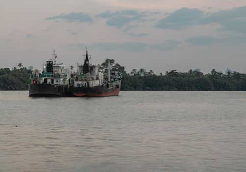 Bangkok, Thailand - 04 Jan 2021 : Two cargo ship parked in the middle of the Chao Phraya River at evening with green forest and sky at the background. Selective focus.