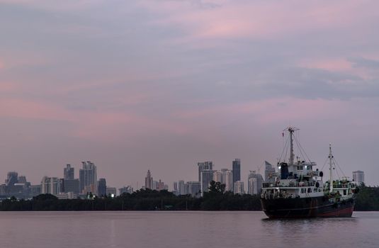 Bangkok, Thailand - 04 Jan 2021 : A cargo ship parked in the middle of the Chao Phraya River at evening with green forest and skyscrapers at the background. Selective focus.