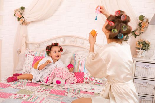 A charming little girl with her mother in hair curlers play with soap balls. Women's day. The girl has fun with her mom.