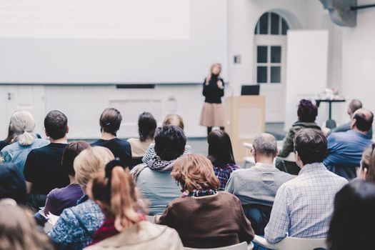 Business and entrepreneurship symposium. Female speaker giving a talk at business meeting. Audience in conference hall. Rear view of unrecognized participant in audience. Copy space on whitescreen.