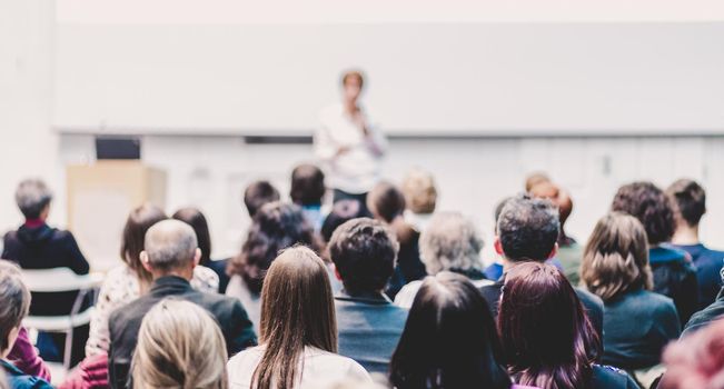 Business and entrepreneurship symposium. Female speaker giving a talk at business meeting. Audience in conference hall. Rear view of unrecognized participant in audience. Copy space on whitescreen.